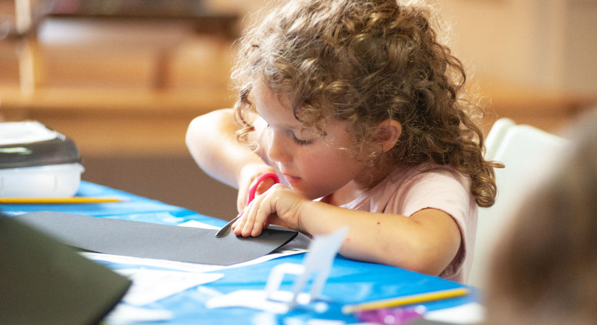 Girl sitting at a desk concentrating while cutting a piece of gray paper with pink scissors. 